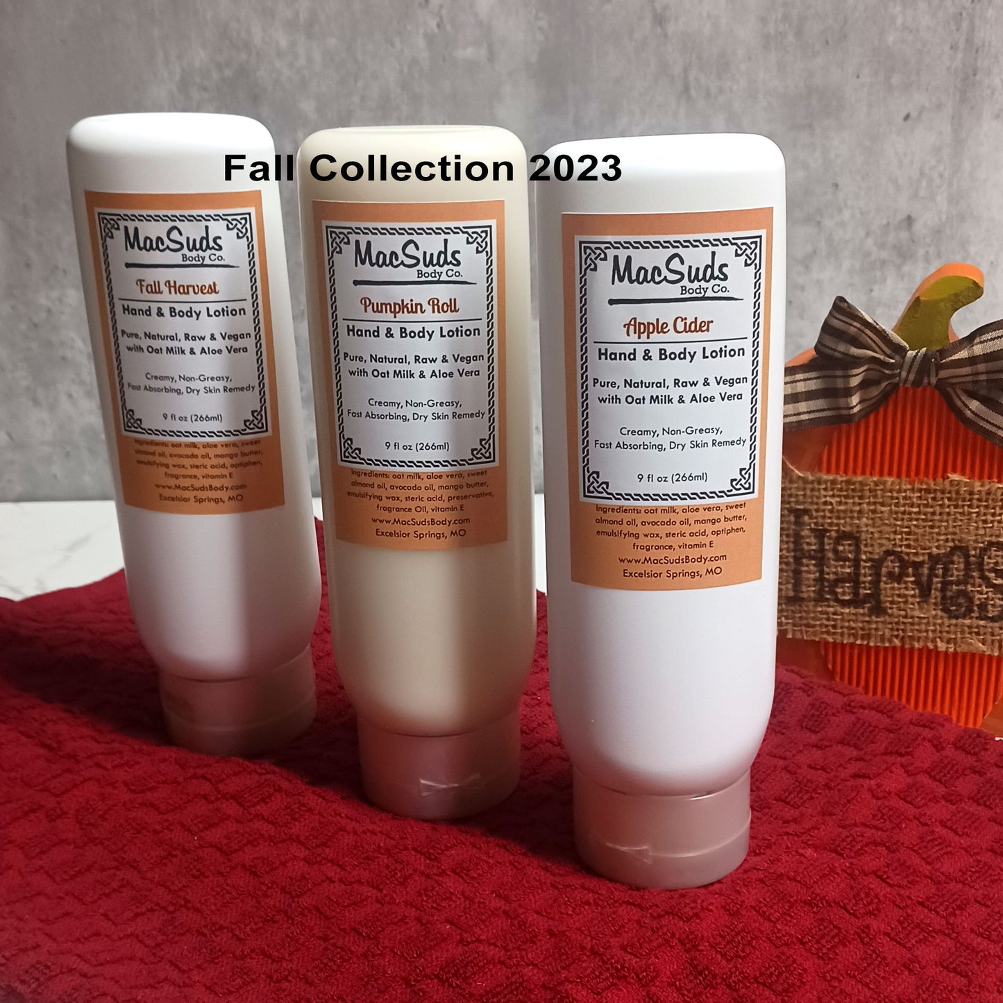 May include: Three white lotion bottles with orange and brown labels. The labels say 'MacSuds Body Co.' and have the names 'Fall Harvest', 'Pumpkin Roll', and 'Apple Cider'. The bottles are on a red surface with a brown and orange fall harvest decoration in the background. The text on the bottles reads 'Hand & Body Lotion', 'Pure, Natural, Raw & Vegan with Oat Milk & Aloe Vera', 'Creamy, Non-Greasy, Fast Absorbing, Dry Skin Remedy', '9